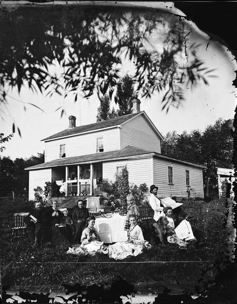 Family posing outdoors around a table in front of a frame house. There are two men with newspapers, girls with watermelon, and a woman on the porch of the house. The Skavlem family is around the table at the home of Lars H. Skavlem. A cloth, books, a vase of flowers and fruit are on the table. The two men on the right have pipes. One has the "Combination Atlas Map of Rock County" (1873); the other has the May 18, 1875 edition of "Skandinaven," a newspaper published in Chicago. The man in the right rear is probably Halvor L. Skavlem. Two women in the front have watermelons. The woman on the front left is probably Halvor's wife, Gunil Olmstad Skavlem. The man and woman on the left are Lars H. Skavlem and Groe Halvorsdatter Aae. Two chairs at the table are empty. A girl is sitting at an organ on the porch, which has many plants on it. A lightning rod is on the roof.
