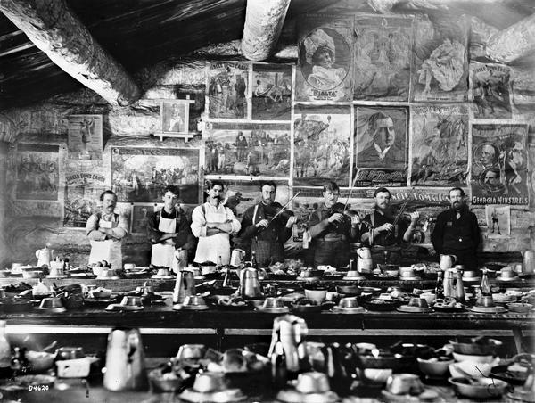 Group of men posing in the dining room at August Mason's lumber camp. Three men are playing stringed instruments. Posters are displayed on the wall behind the men.