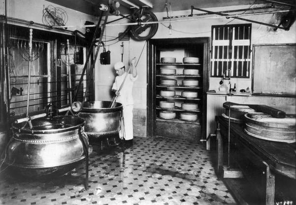 Corner of Swiss cheese-making room, showing a cheesemaker stirring ingredients in a copper kettle.