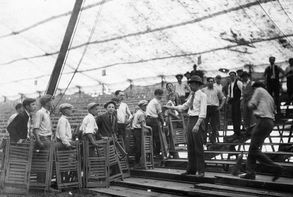 A group of boys, supervised by adult men, set up wooden folding chairs in a circus tent.