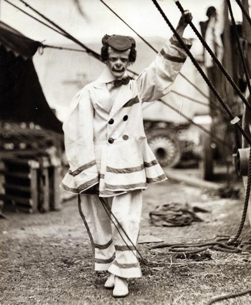 A clown in the Hagenbeck-Wallace Circus standing outside a tent near a wagon, with his left hand holding onto a tent rope. He is wearing a beret-style hat, bow tie, and white shoes.