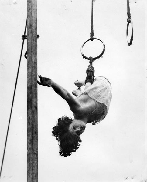 Erma Ward, a circus trapeze artist, dangles by one hand from a ring high in a circus tent.