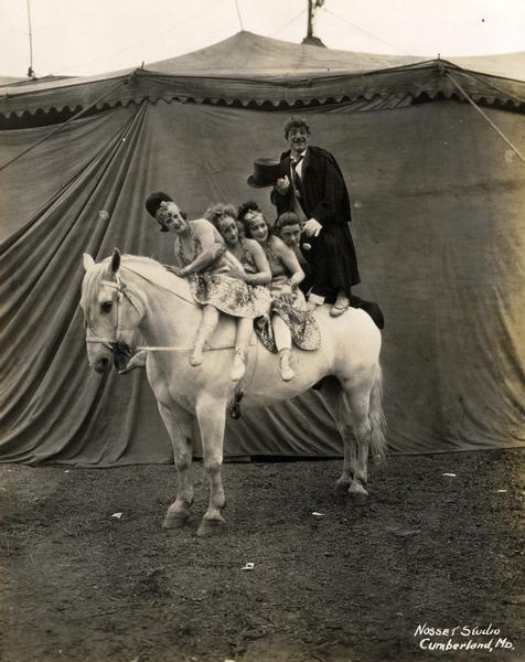 Five circus performers, female and male, sit or stand astride a horse with a tent as a backdrop.