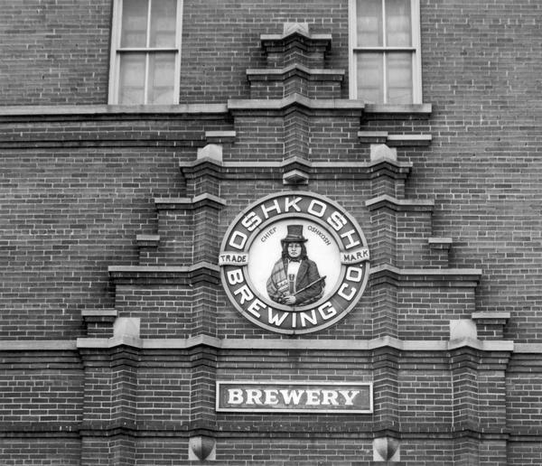 Sign above the main entrance of the Oshkosh Brewing Company, which uses an illustration of Chief Oshkosh as part of its logo.  The sign was installed there when the company opened its new plant in May, 1912.  The sign was removed when the building was demolished in 1986, and is now in the collections of the Oshkosh Public Museum.  This illustration is part of an exhibit about Native Americans created by Paul Vanderbilt, the Wisconsin Historical Society's first curator of photography.