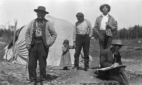 Native Americans on the Menominee Reservation near Keshena, Wisconsin. A white man in the foreground appears to be drawing something out of camera range. This photograph is part of an exhibit about Native Americans created by Paul Vanderbilt, the Wisconsin Historical Society's first curator of photography.