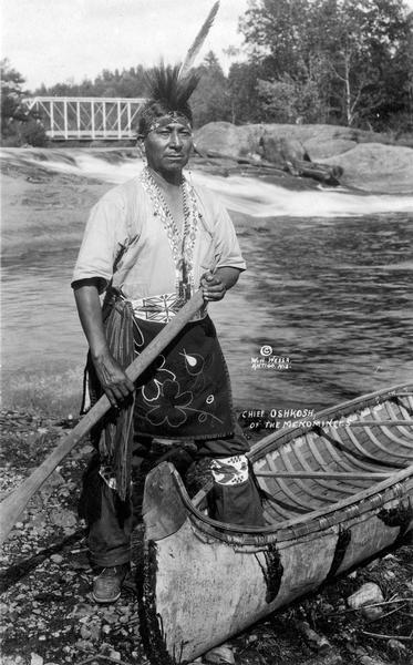 A portrait of a descendant of Chief Oshkosh, probably Ernest Oshkosh, with one foot in a traditional canoe. This image is part of an exhibit about Native Americans prepared by Paul Vanderbilt,  the Wisconsin Historical Society's first curator of photography.  His caption follows:  "Other members of the family of old Oshkosh also achieved prominence.  HIs son  Akwinemi succeeded him as chief, but was also tried for murder in 1871, found guilty and lost his job to his younger brother Neopit Oshkosh, last chief with any authority.  Neopit Oshkosh's sons went away to college.  Photograph of Roy Oshkosh in his capacity as member of the Menominee Indian Advisory Council show him as the most conventionally well-trailer, business-suited member of that group."