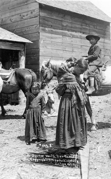 A group of four Native American children thought to have been photographed at the Menominee Reservation. This image is part of an exhibit about Native Americans prepared by Paul Vanderbilt, the Wisconsin Historical Society's first curator of photography. A woman is carrying a child on her back, a young girl stands beside her, and a boy is on horseback behind her. Another horse and a building is in the background.