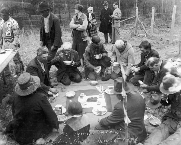 Menominee Indians eating lunch together on Chief Oshkosh Day, May 25, 1926, "the occasion of the removal of the chief's remains from an unmarked grave beside the Wolf River to a spot at the foot of the statue of him by Gaetano Trentanove in Menominee Park in the city of Oshkosh, Wis. A.C. McComb, a public spirited citizen, paid for the transport of the remains and probably provided the lunch. On this day, business was suspended, there was a parade of the Keshena Indian band, with nearly 200 floats, and a banquet at which the Menominee were guests of honor. The crypt of the chief was inscribed, 'Man of Peace, Beloved by All'." This image is part of an exhibit about Native Americans prepared by Paul Vanderbilt.