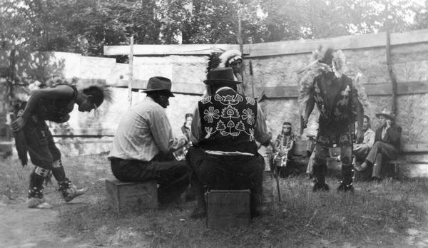 A Menominee ceremonial dance. The dancers are wearing Native American ceremonial dress. Although Roman Catholic missions had banned many Native American ceremonies and dances, rules were gradually relaxed as dances became tourist attractions rather than forms of religious expression. Note the drum circle worn in the grass. This image is part of an exhibit about Native Americans prepared by Paul Vanderbilt, the Wisconsin Historical Society's first curator of photography.