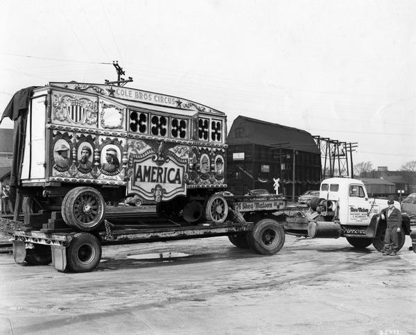 A Cole Brothers Circus wagon sits on a flatbed truck, near a railroad yard, awaiting transport by the Shea-Matson Machinery Mover & Millwrights company.