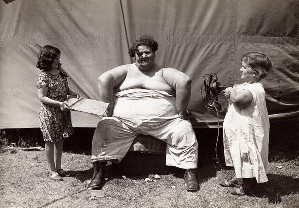 A circus sideshow performer sits outside a tent talking with two women performers of short stature.