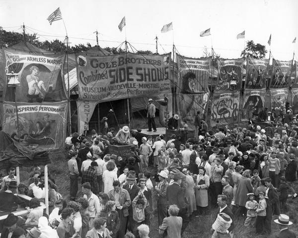 A crowd of circus goers gathers outside the tent of the Cole Brothers Combined Side Shows, with acts announced by a barker standing on a platform.