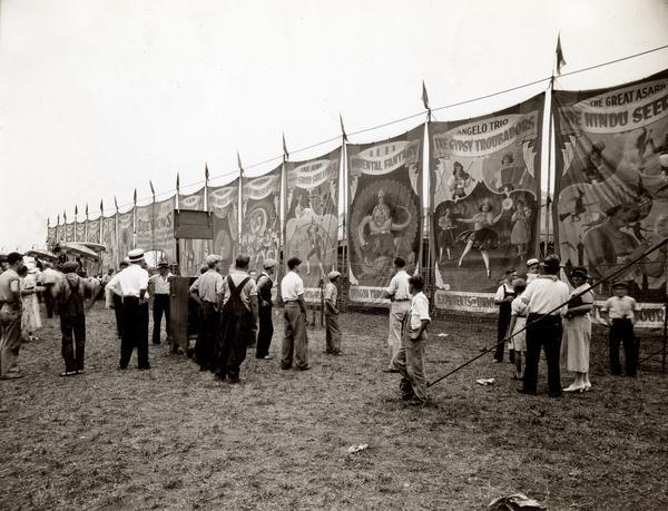 People gather near a circus poster display advertising side show attractions.