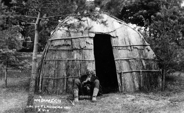 Mr. Bearskin, a medicine man, at Lac du Flambeau sitting in front of of a wigwam covered with elm bark. This image is part of an exhibit about Native Americans prepared by Paul Vanderbilt, first curator of photography at the Wisconsin Historical Society. Caption at bottom left reads: "Lac Du Flambeau Wis."