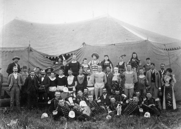 A large group of circus performers and personnel, including a clown and band members, pose in front of a tent.