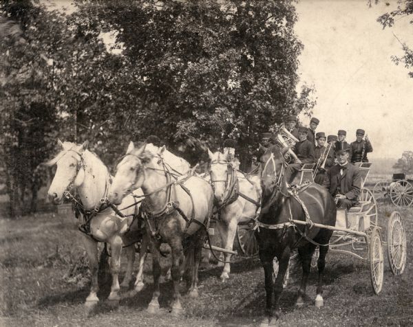 Dode Fisk, the operator of a dog and pony show and circus, drives a wire-wheeled show buggy pulled by the  horse  "Bobby". They are photographed next to a circus bandwagon, with horses and band members.