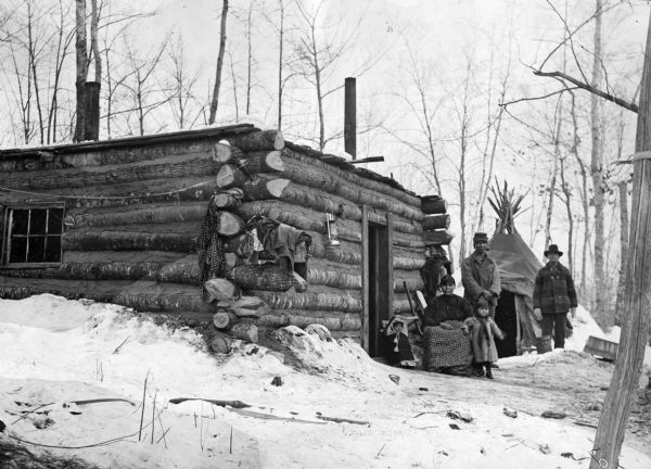 Indians at Big Chetek Lake in Barron County standing near a log cabin and a tipi.  This image is part of an exhibit about Native Americans prepared by Paul Vanderbilt, the first curator of photography at the Wisconsin Historical Society.