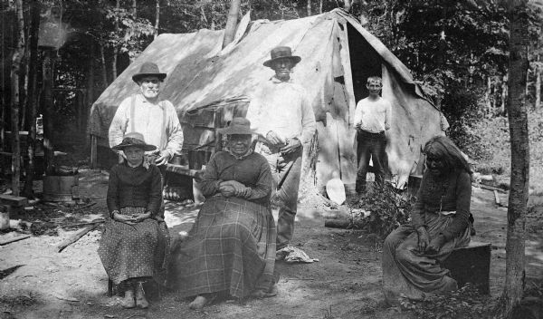 An unexplained group consisting of two white men, two white children, and two Native American women standing in front of a tent.  The original caption stated that the elderly woman to the right was 107-years-old.  This image is part of an exhibit about Native Americans prepared by Paul Vanderbilt, the first curator of photography at the Wisconsin Historical Society.