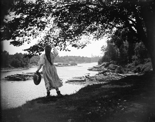 Girl wearing dress, with hat in hand, standing next to a stream. She is shown from the back, looking downstream and waving.