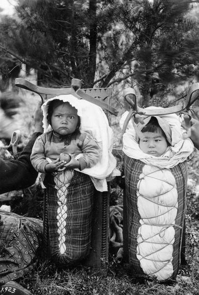 Two Native American infants wrapped in cradleboards. A woman is on the left holding one of the boards, which are propped outdoors in front of trees. This image is part of an exhibit about Native Americans prepared by Paul Vanderbilt, the first curator of photography at the Wisconsin Historical Society.