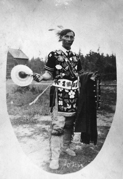 "John," a Native American wearing a combination of traditional and western dress. Among the western items he is wearing are black velvet, sleigh bells, a fan, and a plaid blanket. This image is part of an exhibit about Native Americans prepared by Paul Vanderbilt, the first curator of photography at the Wisconsin Historical Society.
