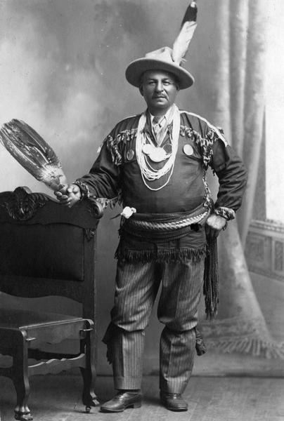 A studio portrait in front of a painted backdrop of an unidentified Chippewa (Ojibwa) Indian from the Bad River Indian Reservation at Odanah. The man is wearing a mix of traditional and western clothing. This image is part of an exhibit about Native Americans prepared by Paul Vanderbilt, the first curator of photography at the Wisconsin Historical Society.