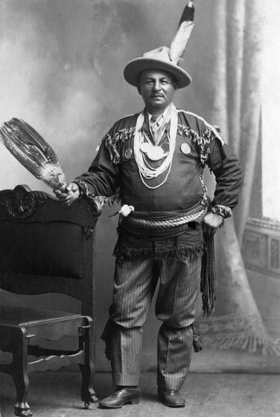 A studio portrait in front of a painted backdrop of an unidentified Chippewa (Ojibwa) Indian from the Bad River Indian Reservation at Odanah. The man is wearing a mix of traditional and western clothing.  This image is part of an exhibit about Native Americans prepared by Paul Vanderbilt, the first curator of photography at the Wisconsin Historical Society.