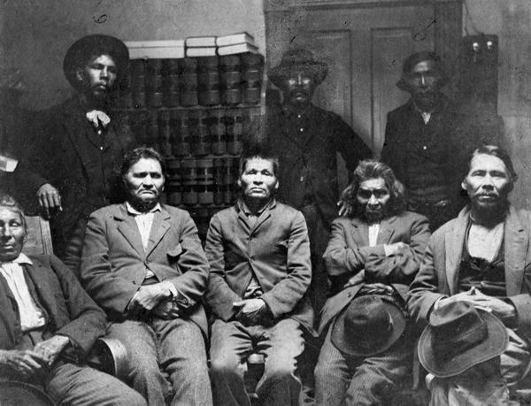 A group of locally important Potawatomi Indians, apparently photographed in a government or law office at Escanaba, Michigan. Left to right standing are Fred Jones Wanka, Joe Negahnee, and Wis-Ki-Ge-Amatyuk, also known as John Buckshot (Prairie Band Potawatomi, Kansas, principle pipe carrier and ritual leader). Seated left to right are Joseph Waumegesake (or Wampum, a descendent of the older chief Wampum, born in Manitowoc), Abraham Michicot (or Mishegand, who was born at Mink River in Door County), David Nsa-waw-quet (or Crutch, reportedly the best hunter and trapper in Michigan), Ben Ah-quee-wee (Dr. Ben), and William Keeshik of the Manitowoc band.