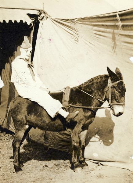 A circus clown sits astride a donkey outside a tent.