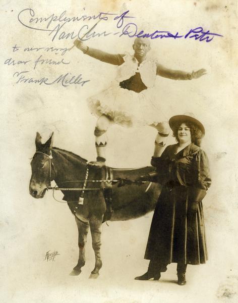 A male circus performer stands lengthwise on the back of a mule, while a female circus performer stands on the ground next to them.