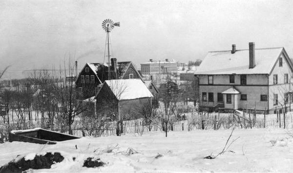 View of University Heights, showing a number of university buildings on the agricultural campus, including a windmill. Lake Mendota is in the far background. Snow is on the ground.