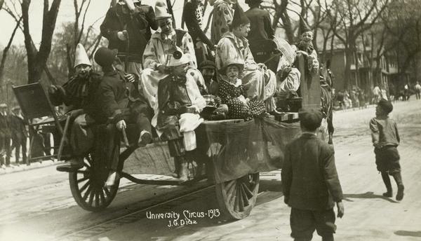 A horse-drawn wagon, filled with University of Wisconsin students dressed as clowns, proceeds along a Madison street as part of a parade. Spectators line the side of the street, while two young boys approach the wagon.