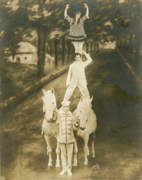 Two horses stand next to each other in front of what may be a painted backdrop of a tree-lined street. A male circus performer stands in the center holding the reins of the horses, while Oscar Lowande, another male performer, stands  on top of the horses, with one leg positioned on the back of each horse. His wife, Mamie Lowande, is standing on his shoulders, creating a pyramid effect. They are members of the Forepaugh & Sells Bros. Circus.