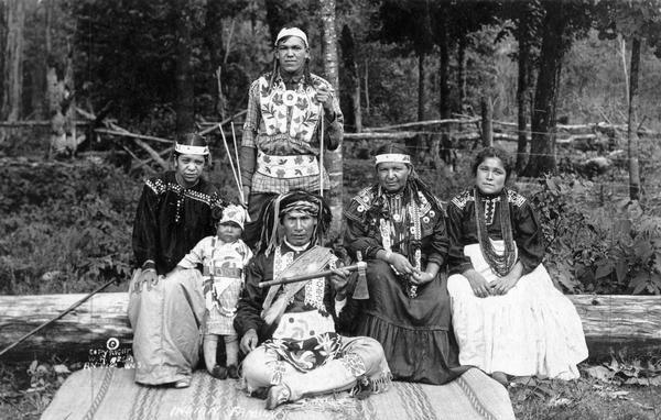 Tecumsa, a Potowatomi, and his family. Tecumsa is standing in the back in the center, and a man is sitting in front of him on a blanket holding up an hatchet. On the left is a woman sitting on a log with a child standing next to her, and two women are sitting on the log on the right. Tecumsa was the keeper of the sacred tribal drum. In the background are fences and trees.