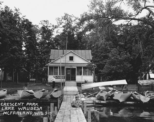 A young boy sits on the dock in front of Olson's store in Crescent Park on Lake Waubesa. Small row boats are arrayed for rental.