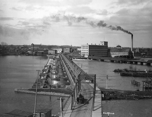 Elevated view of bridge crossing the wide Fox River, with the paper mill on the far side of the river, and a horse-drawn cart approaching the near side.