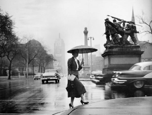 A woman in business attire walking with an open umbrella on a rainy day in downtown Milwaukee. To her left is the Civil War monument, "The Victorious Charge."