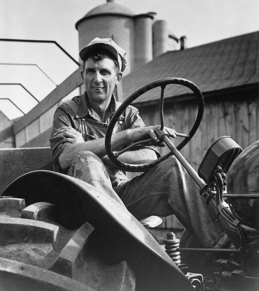 A farmer smiling as he takes the wheel of his tractor. Behind him is a barn and silos.