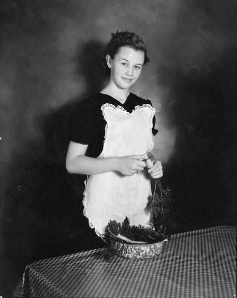 A young woman wearing an apron smiles as she slices a carrot into a pan.