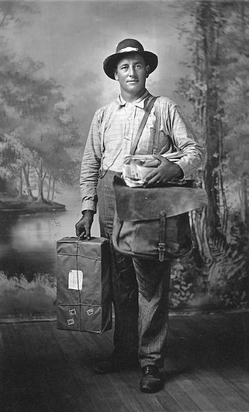 Full-length studio portrait of mail carrier Michael P. Adler, with his mailbag, an armful of mail, and a parcel. He is standing in front of a painted backdrop.