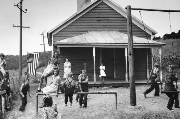 Children play on a playground while a teacher watches from the porch of a one-room schoolhouse. The school building is still located in Grant County, on Highway 61 about 2 1/2 miles south of Boscobel.