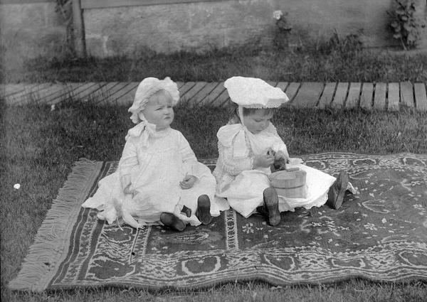 Two small girls dressed in ruffles and bows sit on a woven carpet outside in the sun.