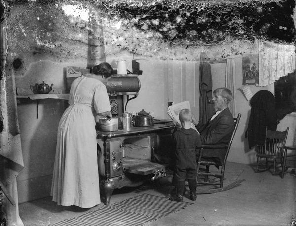 Mr. Peterson is sitting in a rocking chair reading the newspaper and smoking a pipe, while Mrs. Peterson is standing at the stove with their young son, James, nearby.