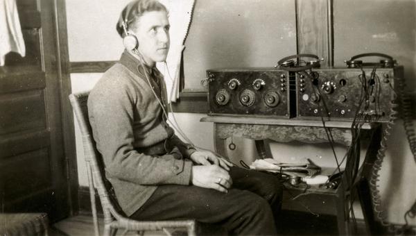 Man seated next to radio set wearing headset.