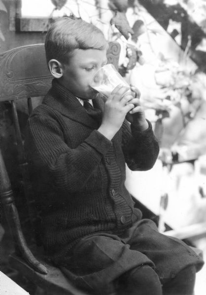 Young boy sitting outdoors on a chair drinking a glass of milk.