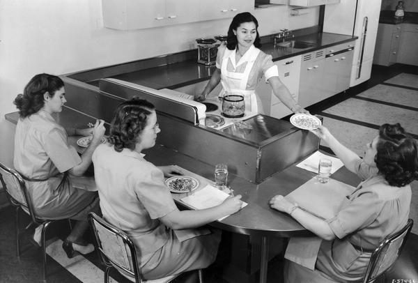 Elevated view of three women evaluating meal prepared by a cook inside International Harvester's Evansville Works home economics kitchen. The Evansville Works produced refrigerators, air conditioners and freezers. Standing behind the counter and serving the food portions is Mrs. Ethel Jean Mitchell. Seated at the counter, from left to right, is: Priscilla Cobb, Ruth Whiting, and Zelma Purchase.