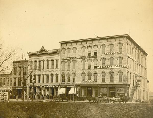 View across Capitol Park lawn towards the corner of West Main and South Carroll Streets with businesses and horse-drawn wagons. The building at the corner is the Fox Block built in 1856, renamed Young's Block, which contains the Worthington & Warner's Business College (became Northwestern Business College, then Capital City Community College) and Ramsay & Campbell Hardware Store.