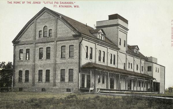 View towards the factory, which has a roof over the long loading dock. Caption reads: "The Home of the Jones' 'Little Pig Sausages,' Ft. Atkinson, Wis.