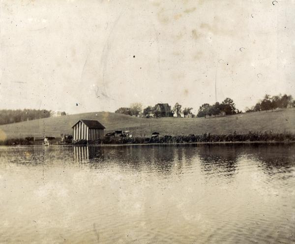 View of shoreline along Picnic Point on the University of Wisconsin-Madison campus showing the home of Professor William Daniels, head of the Chemistry Department. There is a large, striped building on the shoreline, with cows nearby.