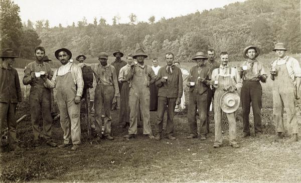 Group of farm workers, men and women, posing in a field, holding various beverages. One man has his arm over a barrel on a stand in the center. In the background is a tree-lined ridge.